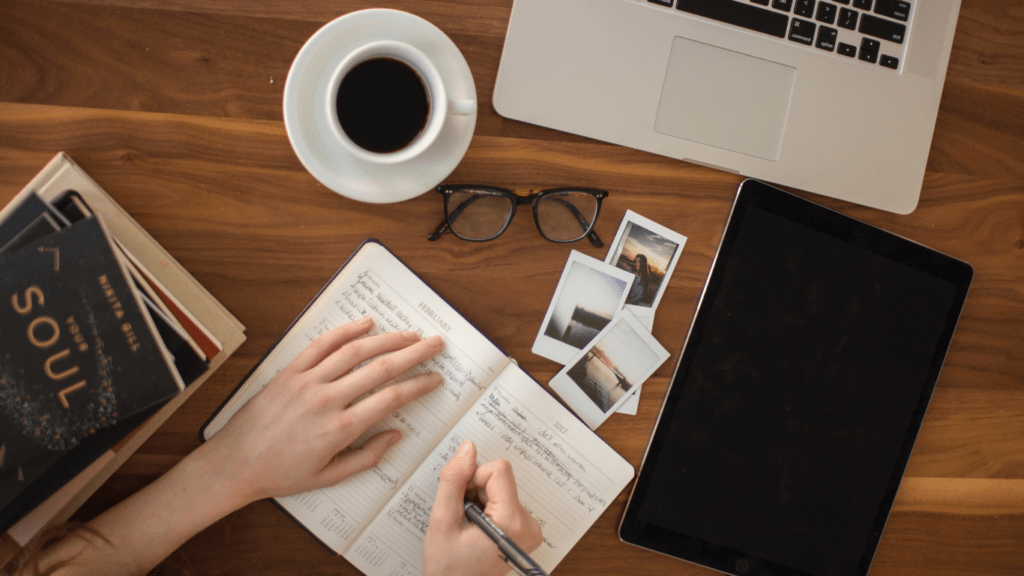 hands writing in a notebook on a wooden table with a laptop